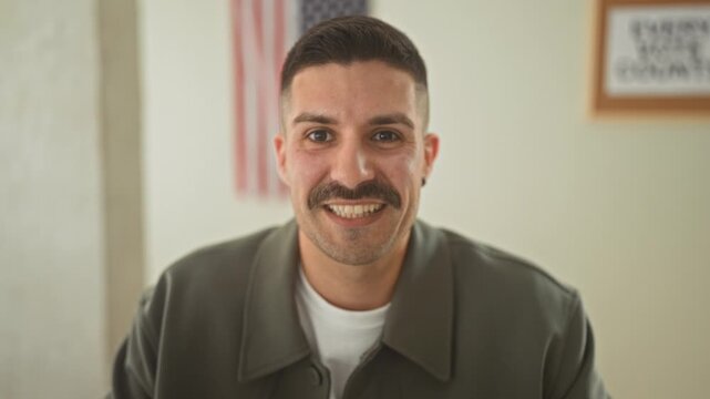 Young man smiles showing teeth with american flag behind him in a polling station building, facing camera and signaling vote support; civic pride.
