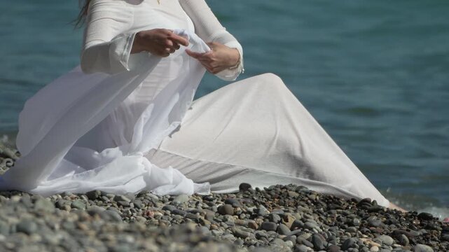 Woman sitting, wearing white dress on rocky shoreline, gracefully waves thin fabric against ocean. Static medium close-up shot.