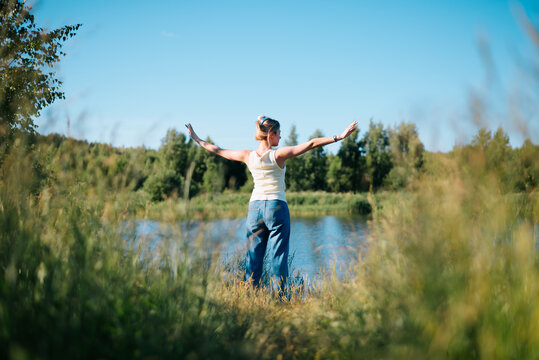 A woman stands with her arms raised in joy by a water body surrounded by greenery. She enjoys the sunny day while appreciating the natural environment around her