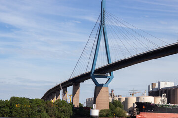 Naklejka premium Modern cable-stayed bridge with blue support towers spanning over a river, industrial buildings and greenery visible along the riverbank