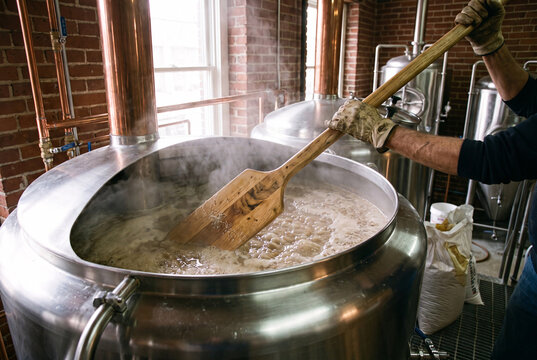 hands holding a large wooden paddle, stirring a massive stainless steel mash tun vat in a craft microbrewery