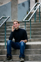 Young man sitting on outdoor stone steps talking on a smartphone smiling during a casual conversation in an urban setting © De Visu