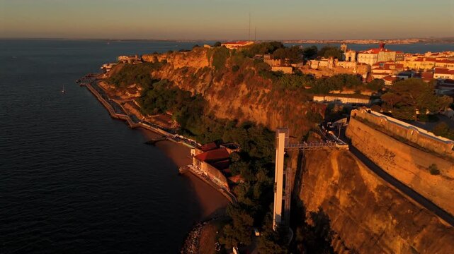 The drone glides alongside the richly textured Boca do Vento cliff revealing warm golden rock beneath a town at dusk.