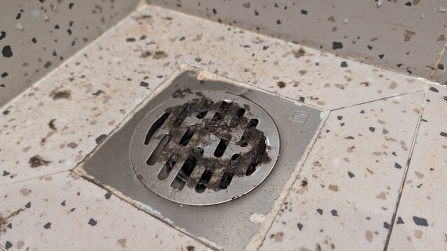 Close-up of a dirty and clogged shower drain on a tiled floor, full of dust, dirt, and hair. Concept of household maintenance, cleaning, and plumbing issues in the bathroom.