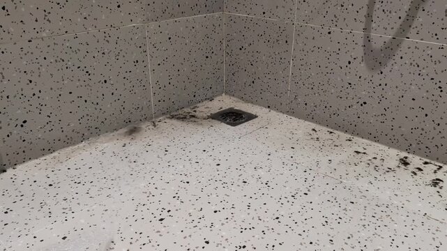 View of a dusty and dirty tiled shower floor being rinsed by a stream of water, creating a slow drainage effect as grime clogs the drain.