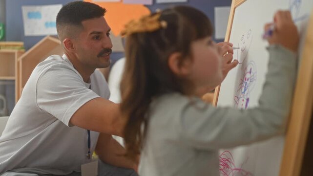 Young girl holds marker drawing on easel while male teacher watches and guides hand in school building; curiosity learning.
