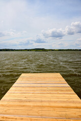 Obraz premium Lutskovlyany lake reservoir near Hrodna, Belarus. Wooden pier leads into calm water under cloudy sky. Peaceful summer nature landscape shows rural beauty, ripples on surface, green shoreline far away.