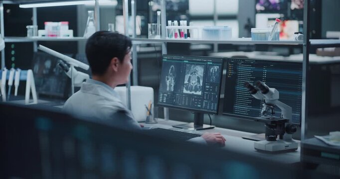 Asian Male Researcher walks toward his Workspace, sits down to  Study Neurological Brain Scan Imagery on Screens at a Desk in an Medical Laboratory.Pharmaceutical Drug Development, Disease Treatment