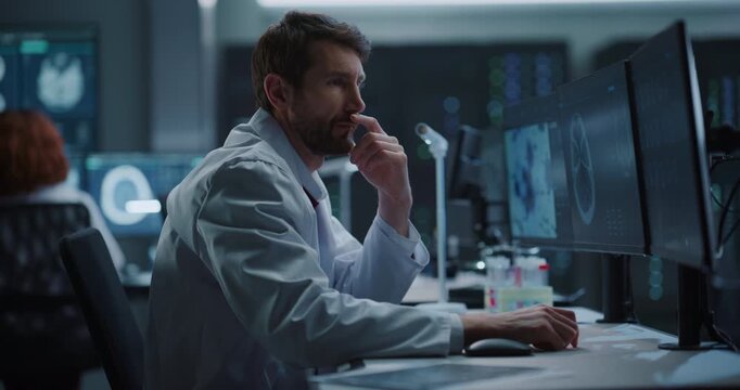 Male Scientist Analyzes Brain MRI Scans on Dual Computer Monitors in a Modern Medical Research Laboratory, Applying AI Technology for Neurology Diagnosis, Drug Development and Disease Treatment.