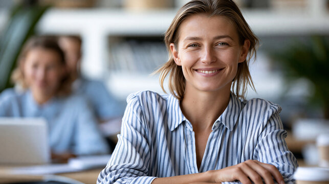 Faceless smiling woman resting hand on desk glancing toward camera while colleague works quietly in softly blurred background, collaborative office concept, corporate photography,