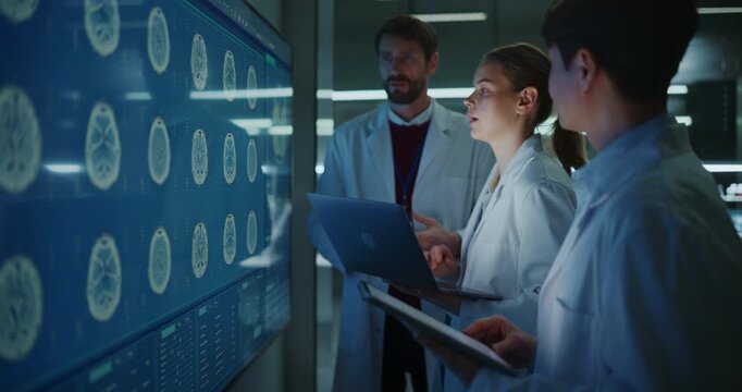 Multiethnic Group of Neurologists Talk and Point at MRI Brain Scans on a Large Screen, Holding Laptop and Tablet During a Meeting in a Modern Research Hospital Laboratory Developing Neurology Therapy.