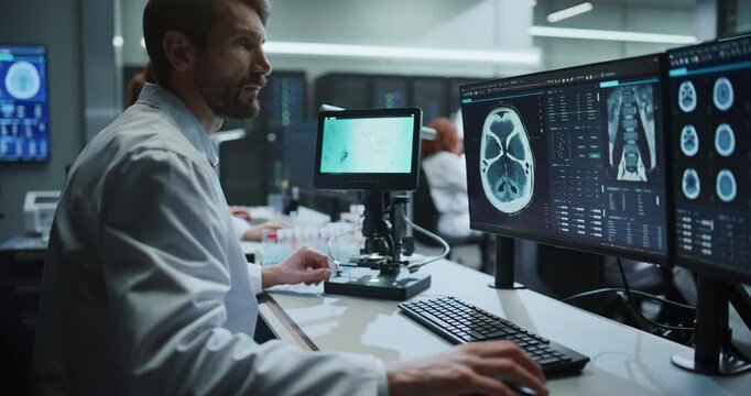 Male Scientist Uses Digital Microscope to Analyze Samples While Referencing MRI Brain Scans on Screens in Modern Medical Research Hospital, Colleagues Work on Drug Development for Disease Treatment