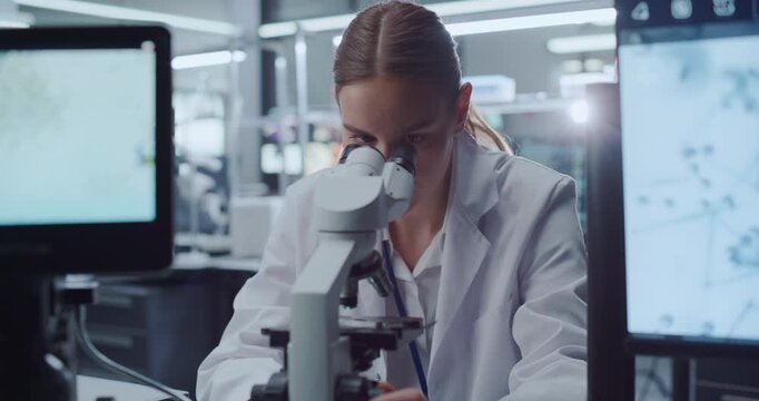 Caucasian Female Scientist Looks into Microscope Examining Molecular Samples While Data Displays on Monitors in Advanced Hospital Laboratory as Colleagues Pursue Drug Development for Disease Cures