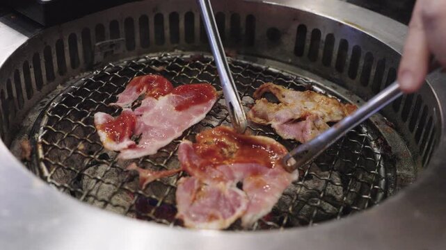 Close-up of thin marinated meat slices grilling on a metal round grate over charcoal. Tongs are flipping the meat in a Korean BBQ or Japanese Yakiniku restaurant. Delicious grilled food concept.