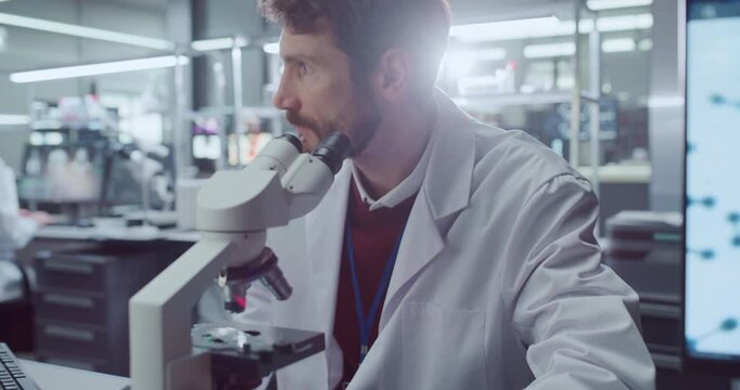 Portrait of Male Scientist Peers Through Microscope Examining Biological Samples in Medical Research Hospital Laboratory While Colleagues Reviews Data Screens for Drug Development. Cinematic Close-up
