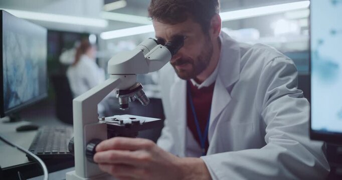 Portrait of Male Physician Examines Biological Specimens Through Microscope in Medical Hospital Laboratory as Colleagues Analyzes Molecular Data on Screens Disease Treatment Research. Cinematic Camera