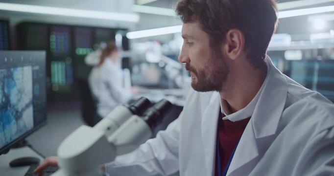 Portrait of Male Researcher Examines Biological Samples Looking Under Microscope, Studies Molecular Imaging on Screens in an Advanced Medical Hospital Laboratory For Research Disease Treatment