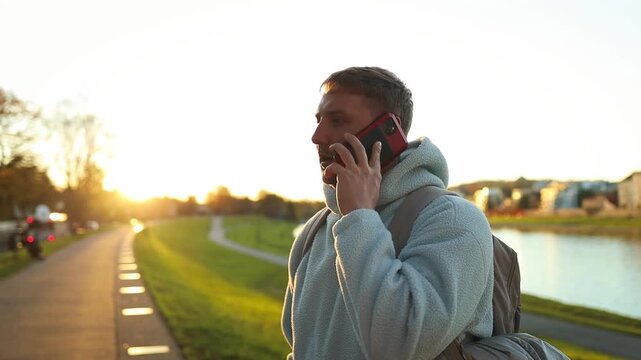 A man is casually walking and talking on the phone along a concrete pathway near a water surface during sunset. The scene conveys a relaxed and casual mood, reflecting daily life and social.