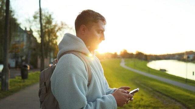 A man is casually using his smartphone while walking through a park-like setting with trees and grass. The scene captures a relaxed and urban lifestyle, emphasizing technology use in public.