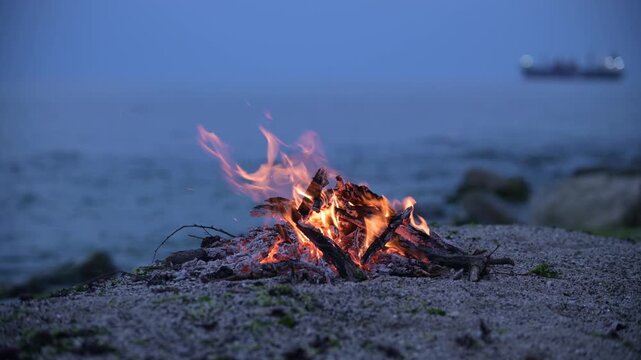 Bonfire burning on a rocky beach at dusk with cargo ship on the horizon