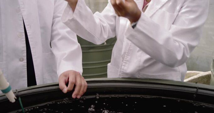 Two lab techs in coats, probe dipping into tank in greenhouse, woman sampling cup testing water