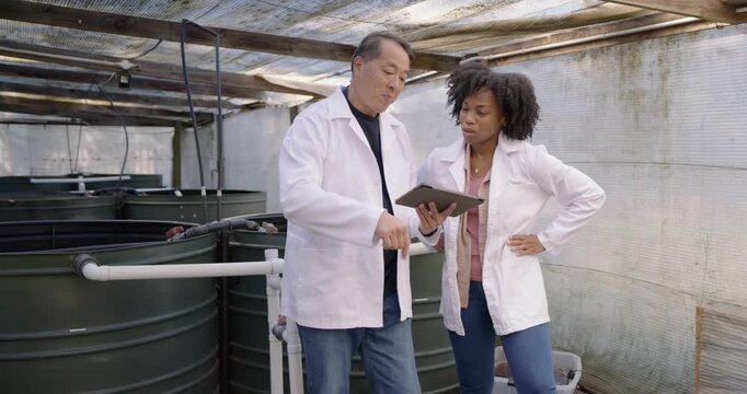 Diverse coworkers in lab coats inspecting tanks and piping in greenhouse after checking tablet