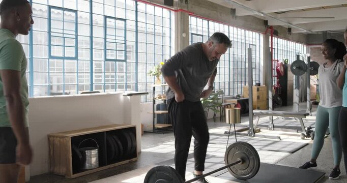 Diverse group clapping, coach prepping, performing clean press with loaded barbell in gym, showing