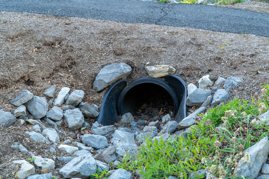 Black plastic culvert drain pipe installed in a rocky, mulched embankment beside an asphalt road, providing essential drainage in a natural outdoor setting.