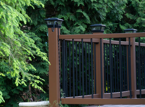 Modern brown composite deck railing with sleek black balusters and solar post cap lights, set against a vibrant green evergreen hedge, enhancing outdoor living and home exterior aesthetics.