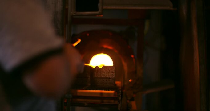 A craftsman working with molten glass in a furnace in a dimly lit studio