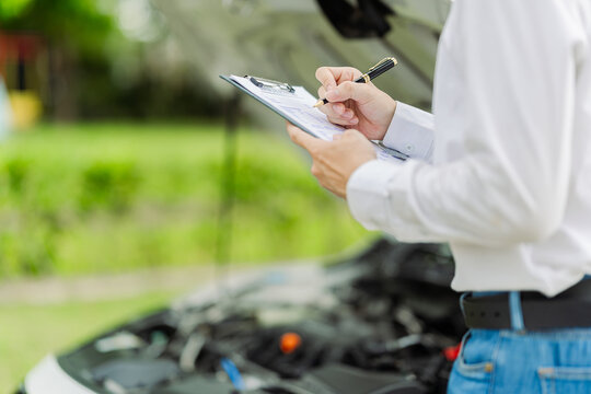  Car insurance agent inspecting engine under open hood and taking notes on clipboard. Professional technician or adjuster checking vehicle engine condition after accident.