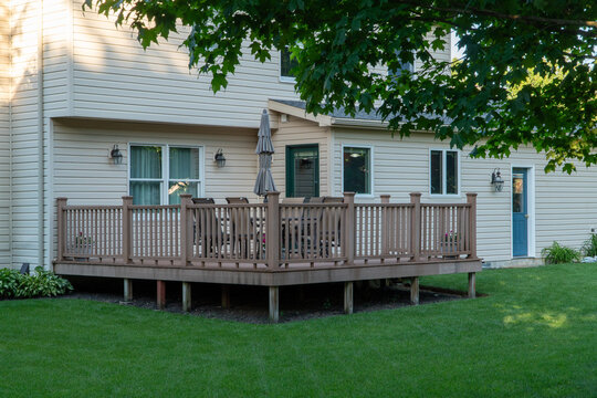 Modern suburban home with spacious composite deck, outdoor dining set, and closed umbrella, surrounded by lush green lawn. Features beige vinyl siding, multiple windows, and a vibrant green backyard.