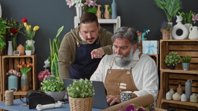 Two men florists in aprons lean over a laptop, one points finger to screen and gestures at online order while the older bearded florist holds a mug in a flower studio; focused teamwork.