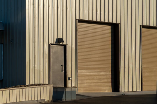 Industrial building exterior with beige corrugated metal siding, a dark loading dock access door, and a large brown roll-up garage door, bathed in warm sunlight creating dramatic shadows.