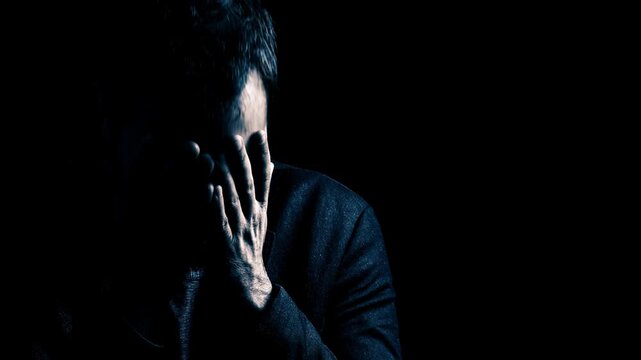 Desperate man covering face with hands in darkness. Depressed guy suffering from extreme burnout and mental struggle. Emotional breakdown and psychological distress.