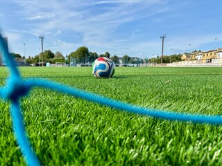 Soccer ball on artificial green turf with goal net in foreground under blue summer sky © Iryna
