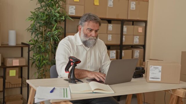 Man scans a cardboard package with a handheld barcode scanner as colleague holds the box near a laptop in a small business warehouse building full of stacked boxes; teamwork efficiency.