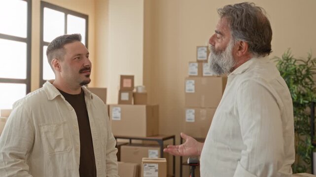 Two men with stacked cardboard packages stand smiling with arms crossed in a building shipping area among labeled boxes and a plant by the window; small business pride teamwork.
