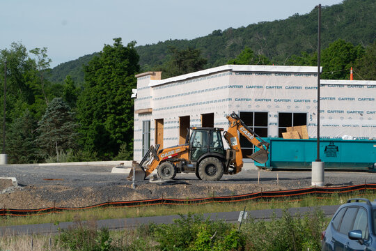 New commercial building under construction with a yellow CASE backhoe loader, large waste dumpster, and erosion control fencing on a sunny day with green hills in the background.