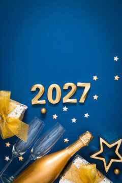 Happy New Year 2027 Vertical Banner with Gold Numbers, Champagne Bottle, Two Glasses and Gift Boxes on Classic Blue Background. Festive Winter Holiday Greeting Card Layout with Copy Space.