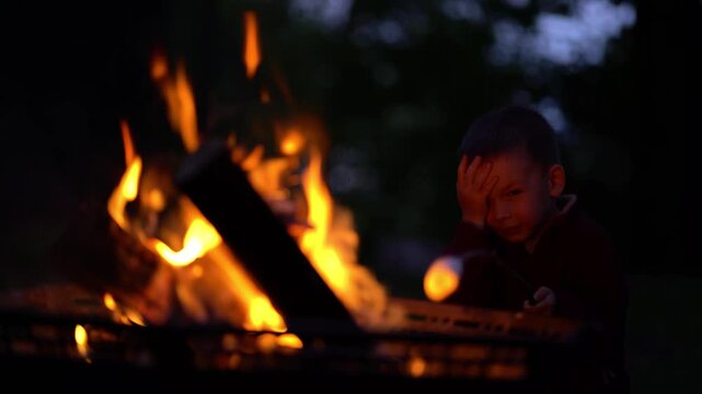 Young boy roasting marshmallow over glowing campfire at night. Focused child in warm firelight during cozy family camping. Perfect summer outdoor leisure