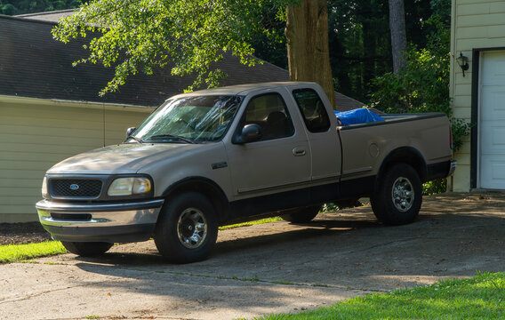 Ford F-250 Super Duty pickup truck parked on a sunny concrete driveway in front of a suburban home with lush green trees and a garage, showcasing a classic American vehicle.