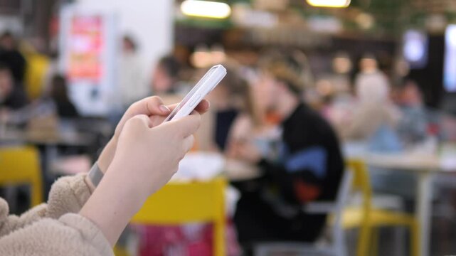 Person holds a smartphone in a busy cafe. Background shows blurred diners at yellow tables. Focus is on hand and device. Scene captures modern digital engagement in casual spaces