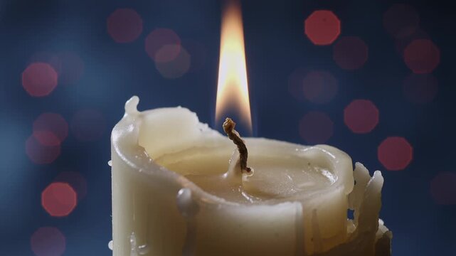 Close-up of a burning white candle with a flickering flame. Melted wax texture against a dark blue background with red bokeh lights