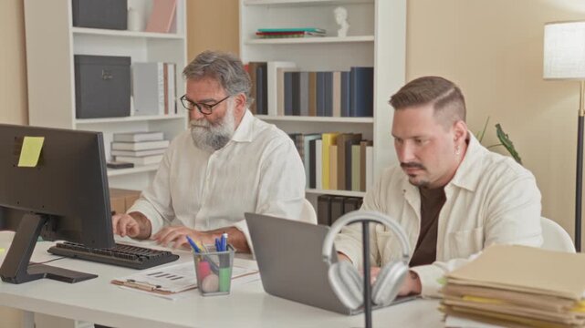 Two men type on a keyboard and flip file folders with visible hands at a desk in building; teamwork concentration.