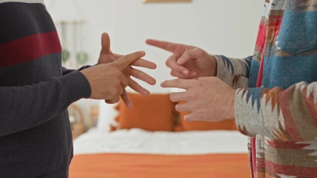 Two men reaching and interlacing hands above a bed in a building bedroom with orange pillows and casual sweaters; intimacy connection tenderness.