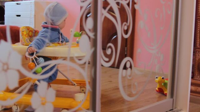 A toddler in a baby walker looks through a glass door, A boy learns to walk with the help of a baby walker