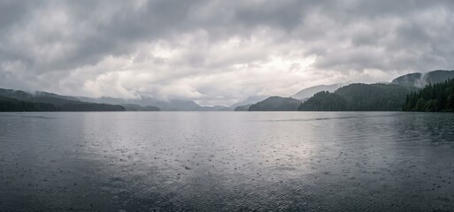 Fototapeta premium Panoramic view of a calm lake under a dramatic overcast sky with rain falling creating ripples on the water's surface and distant misty mountains