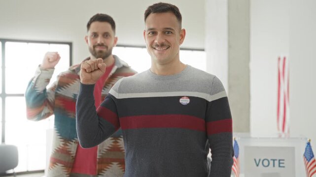Two men raise fists with an i voted sticker at a polling building near a ballot box and us flags, smiling at camera; pride civic duty.