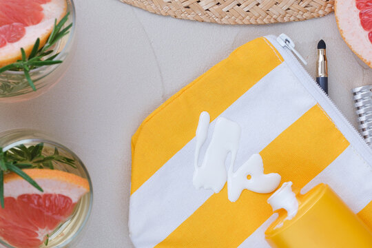 Top view of white sunscreen lotion spilled from a yellow bottle onto a striped beach pouch surrounded by summer accessories, grapefruit, and a refreshing drink.
